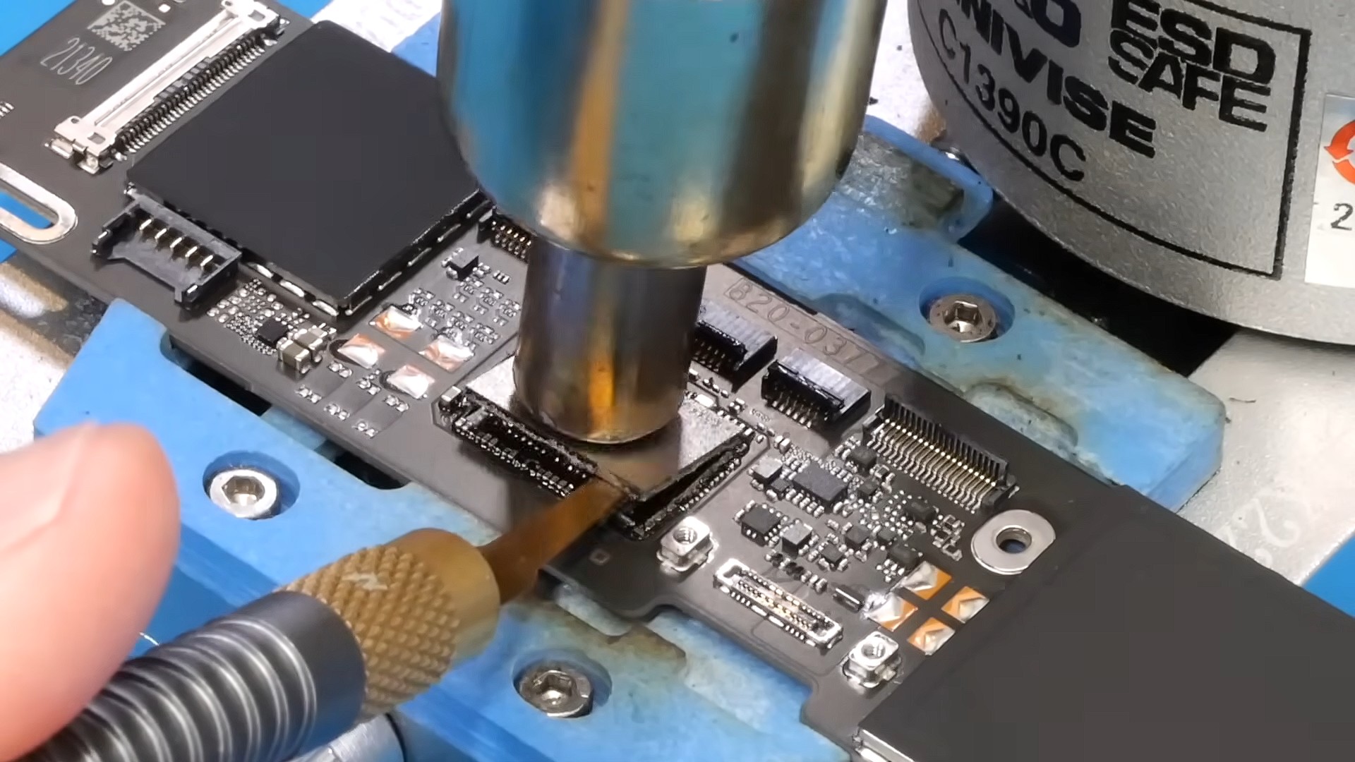 من موقع فون إسلام: شخص يستخدم أداة لحام ومحطة هواء ساخن للعمل على المكونات الإلكترونية الصغيرة أثناء ترقية ماك بوك نيو، مع تثبيت لوحة الدائرة الكهربائية بإحكام في ملزمة زرقاء.