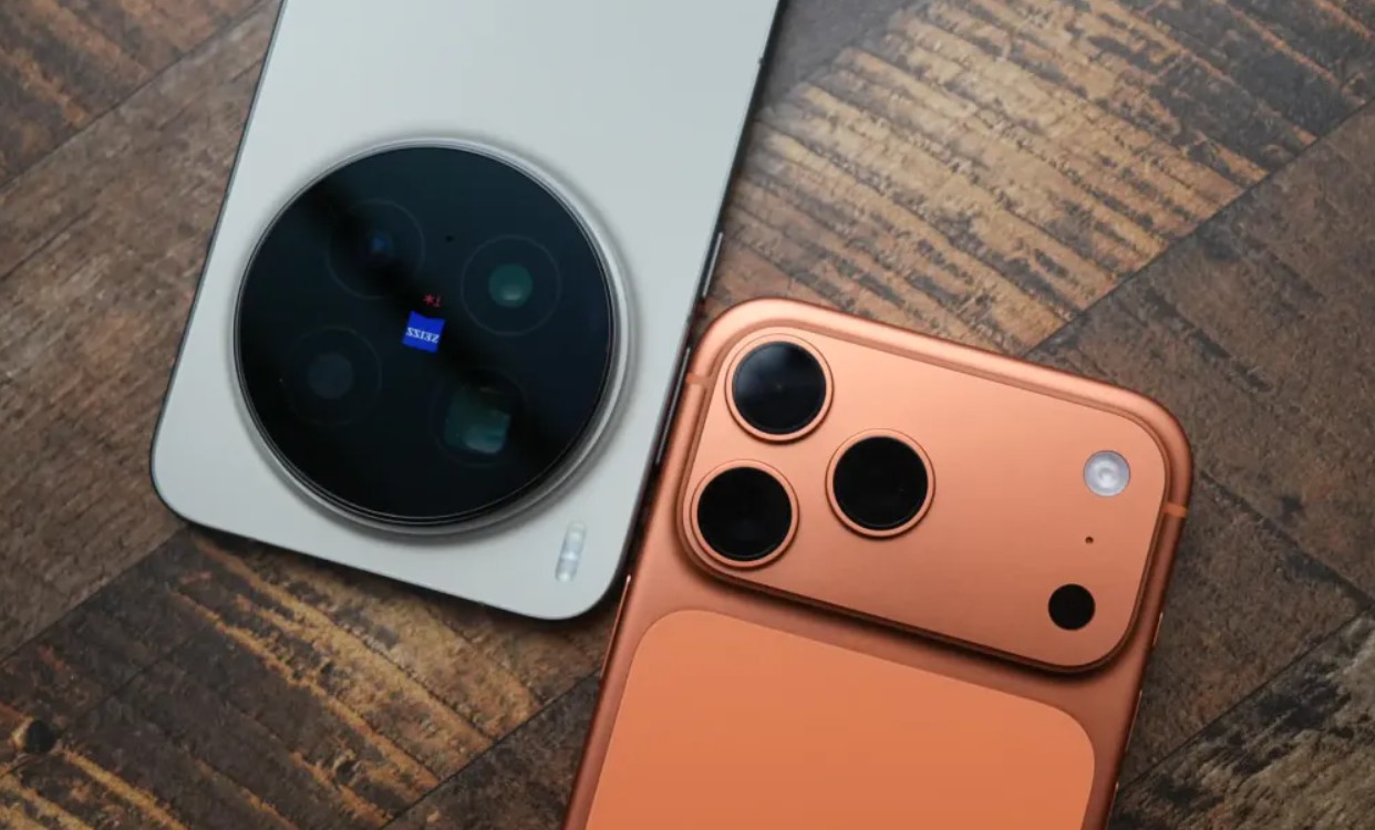 من موقع فون إسلام: يوجد هاتف ذكي باللون البيج الآي-فون ذو كاميرا خلفية دائرية كبيرة باللون البيج مع وحدة كاميرا خلفية برتقالية اللون قابلة للطي مع كاميرا ثلاثية وكلاهما موضوعان على سطح خشبي.