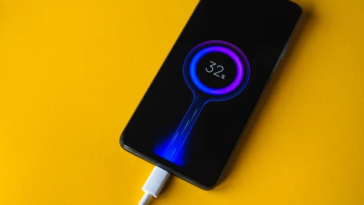 من موقع فون إسلام: هاتف ذكي يشبه الآي فون يتم شحنه بكابل USB أبيض اللون ويظهر على شاشته مستوى بطارية بنسبة 32% على خلفية صفراء نابضة بالحياة.