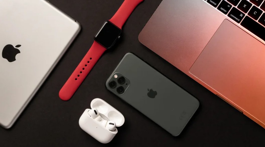 من موقع فون إسلام: تم ترتيب جهاز iPad، وساعة Apple Watch بسوار أحمر، وساعة Apple Watch بسوار أحمر، وساعة آيفون كالهاتف المناسب، وسماعات AirPods Pro في علبة شحن، وجهاز MacBook على سطح أسود.