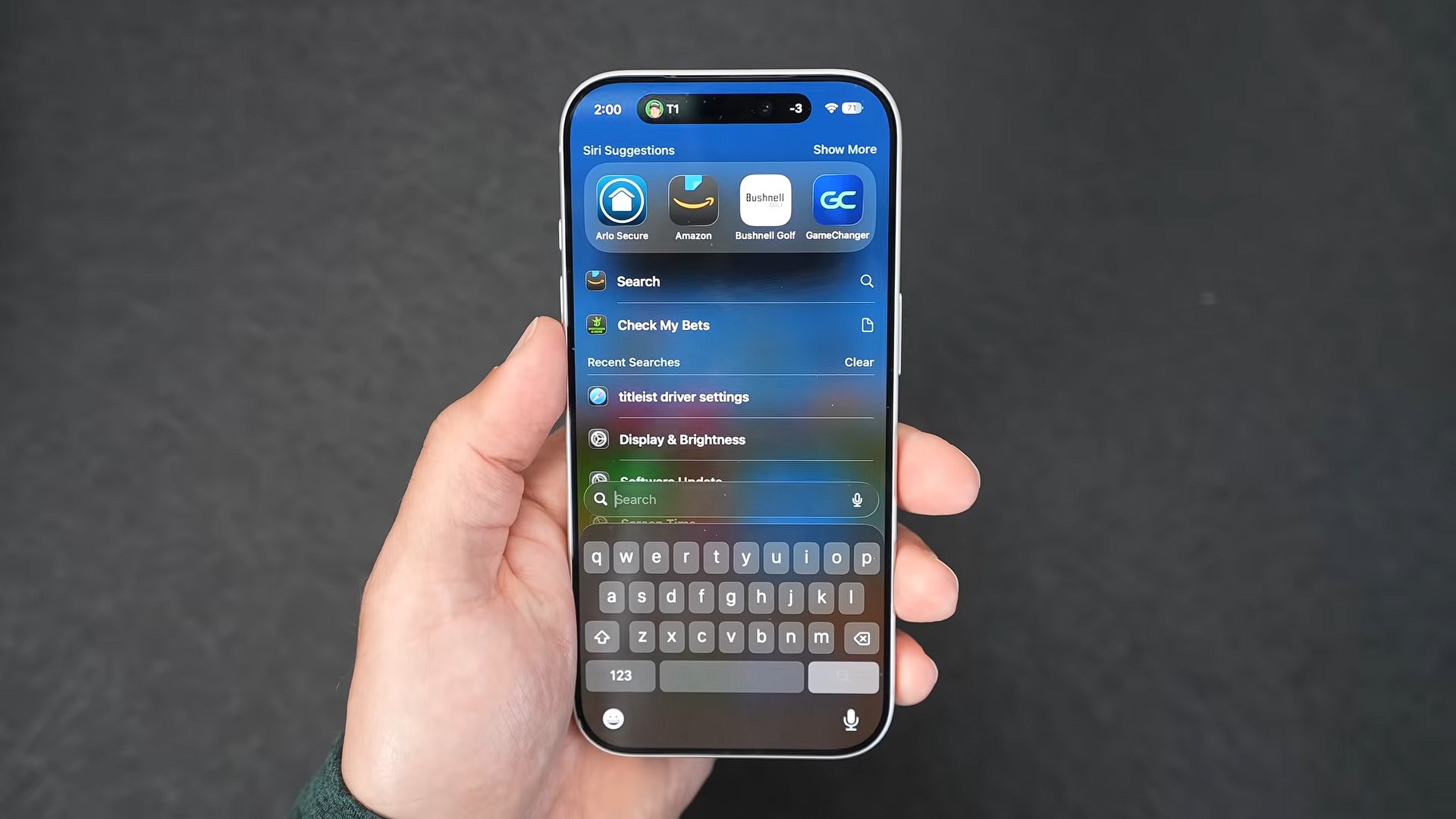 من موقع فون إسلام: يد تحمل جهاز iPhone وشاشة البحث مفتوحة، وتظهر اقتراحات التطبيقات ولوحة مفاتيح معروضة وتظهر سيري في نظام iOS 27 على خلفية داكنة.