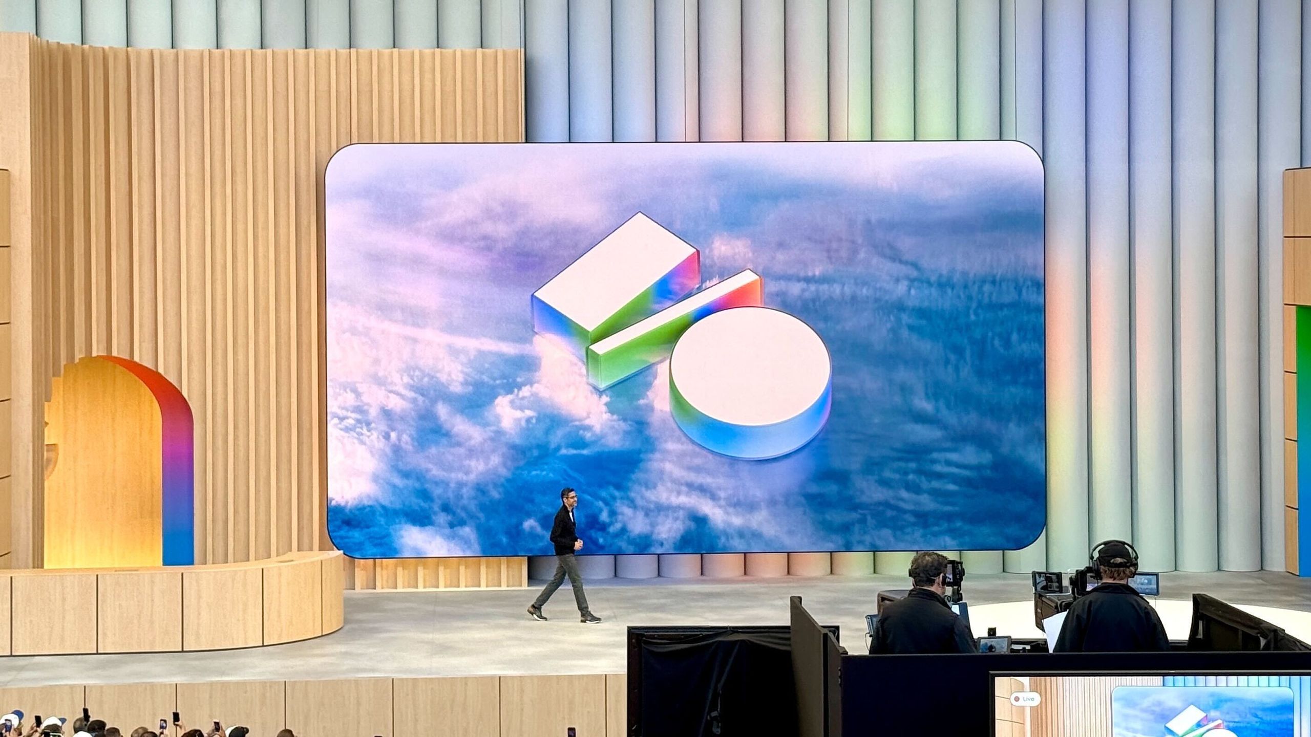 من موقع فون إسلام: شخص يسير على خشبة المسرح أمام شاشة كبيرة تعرض شعار مؤتمر Google I/O 2026، بينما يجلس موظفو الحدث في محطة عمل في المقدمة.