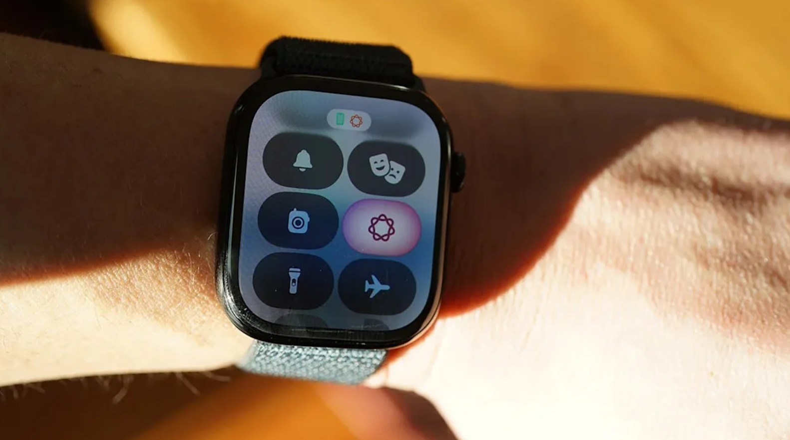من موقع فون إسلام: ساعة ذكية على معصم الشخص، محدثة بنظام تحديث watchOS 27، تعرض مركز التحكم الخاص بها الذي يضم أيقونات لإعدادات مثل الوضع الصامت، ووضع المسرح، والكاميرا، والتركيز، والمصباح اليدوي، ووضع الطائرة.