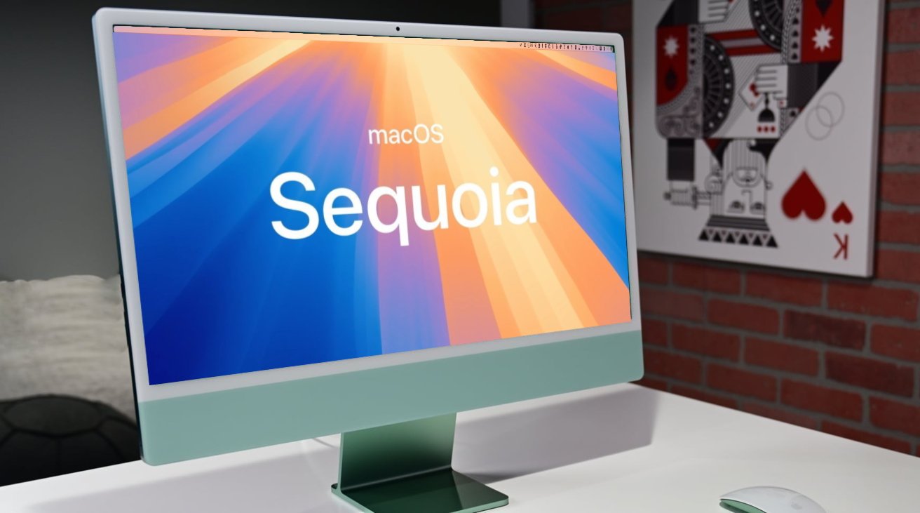 من موقع فون إسلام: يعرض جهاز كمبيوتر iMac على مكتب شاشة بدء تشغيل macOS Sequoia؛ وفي الخلفية جدار من الطوب الأحمر وفن بطاقات اللعب المؤطرة، ويظهر تصميم أنيق ومثالي لعملاء أمازون غلوبال ستارار.