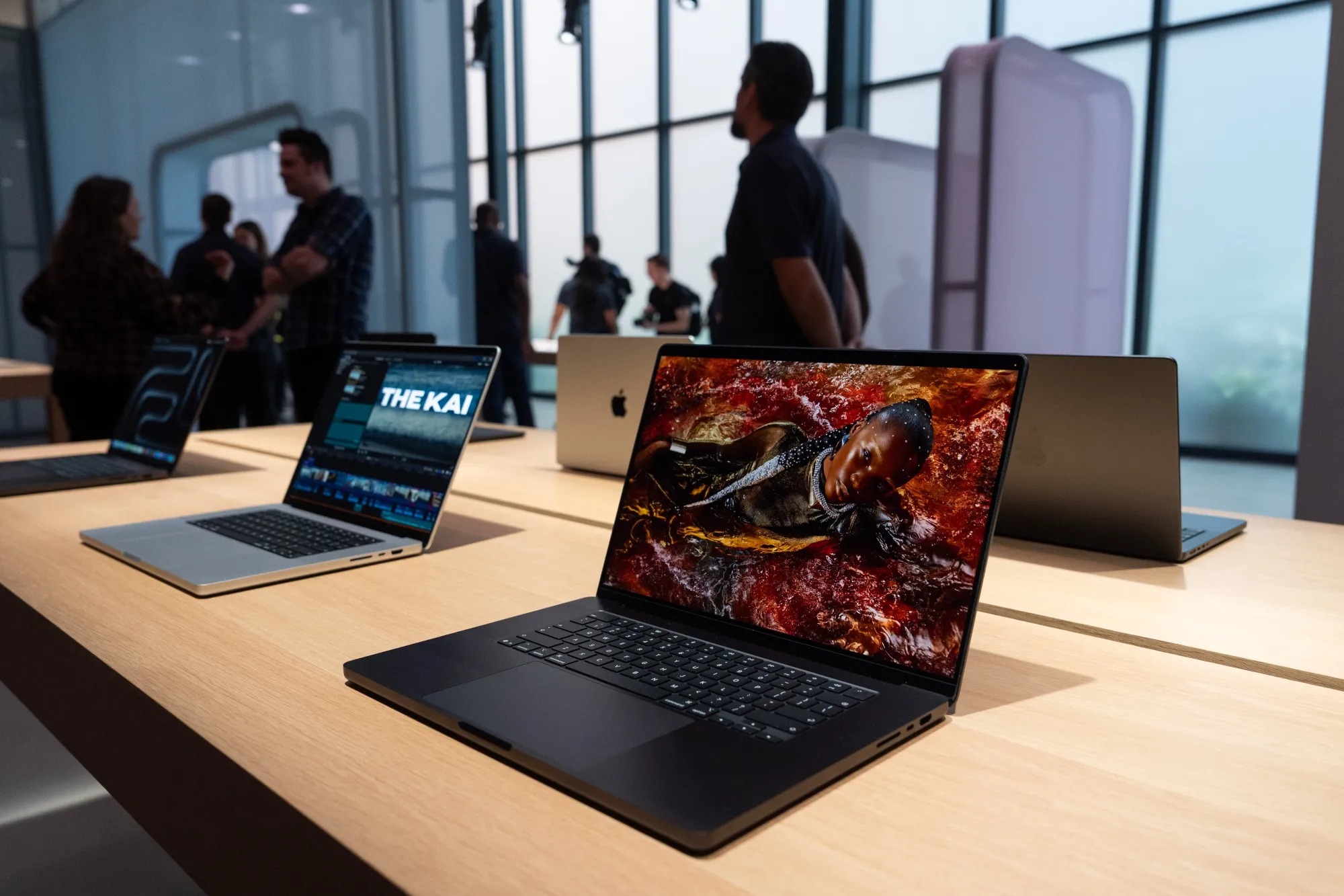 من موقع فون إسلام: تُعرض عدة أجهزة كمبيوتر محمولة من طراز MacBook مفتوحة، مزودة بمعالجات آبل M7، على طاولة خشبية داخل متجر، بينما يقف أشخاص في الخلفية ويتحدثون.