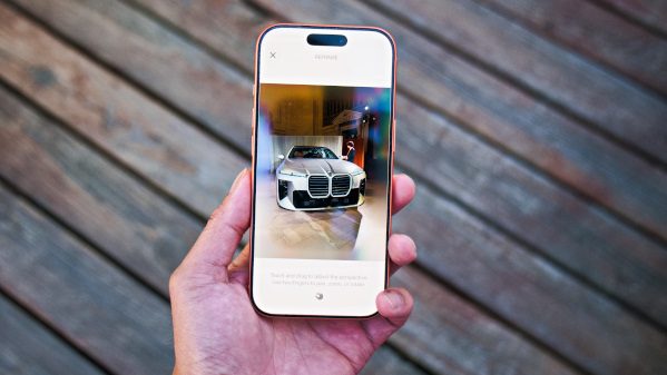 من موقع فون إسلام: تُظهر يد تحمل هاتفًا ذكيًّا يعمل بنظام التشغيل iOS 27 صورة لسيارة BMW بيضاء اللون، مع سطح خشبي في الخلفية.