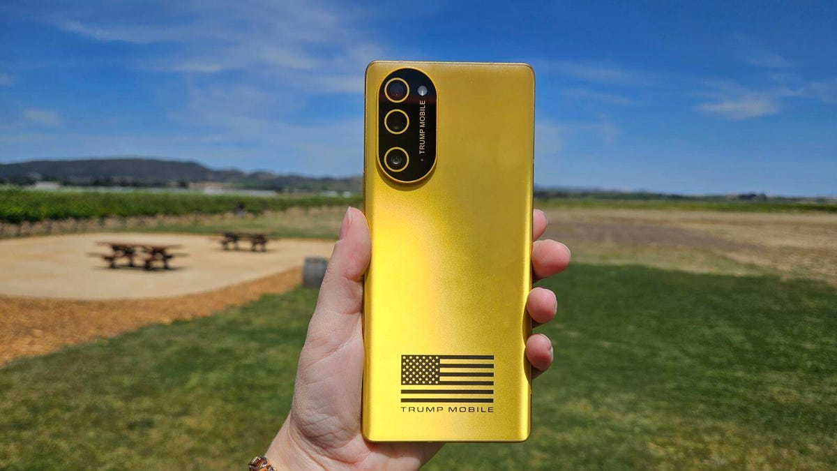 From Phonegram website: A hand holding a gold-colored 'Trump T1' smartphone, featuring the 'TRUMP MOBILE' logo and a black and white American flag on the back, outdoors with grass and a picnic area in the background.