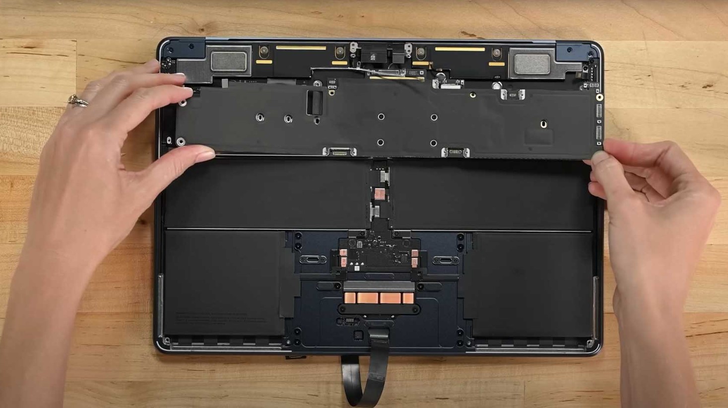 من موقع فون إسلام: يقوم شخص برفع الغطاء الداخلي لجهاز MacBook Air تم تفكيكه جزئيًا على سطح خشبي، ليكشف عن مكوناته الداخلية، بما في ذلك المروحة.