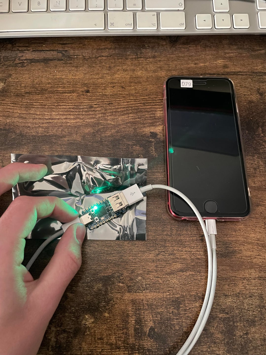 من موقع فون إسلام: يد تمسك بجهاز USB يضيء منه ضوء أخضر، وهو متصل بهاتف ذكي عبر كابل USB على مكتب خشبي تظهر في الخلفية لوحة مفاتيح، وربما يكون صاحب اليد بصدد التحقيق في ثغرة أمنية في جهاز آيفون قديم.