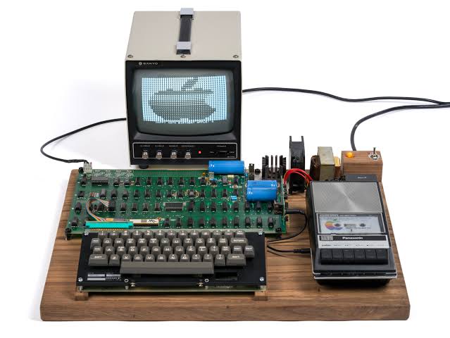 من موقع فون إسلام: إعداد مبكر لجهاز الكمبيوتر «Apple-1» يتضمن لوحة مفاتيح، ولوحة دوائر كهربائية، ومسجل أشرطة كاسيت، ومصدر طاقة، وشاشة تعرض مخططًا لشعار شركة «Apple».