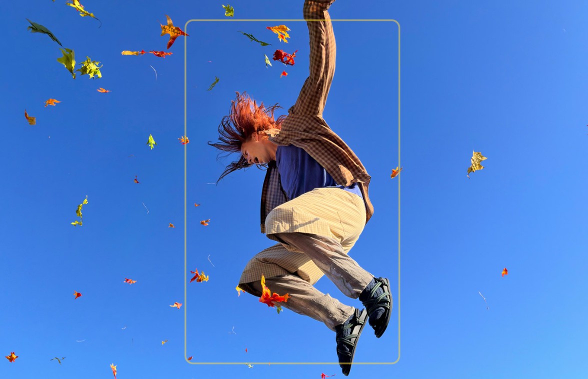 From the Phonegram website: A person wearing beige pants and a blue shirt jumping in the air against a clear blue sky, surrounded by scattered autumn leaves — perfectly capturing this vibrant moment thanks to the iPhone camera features.