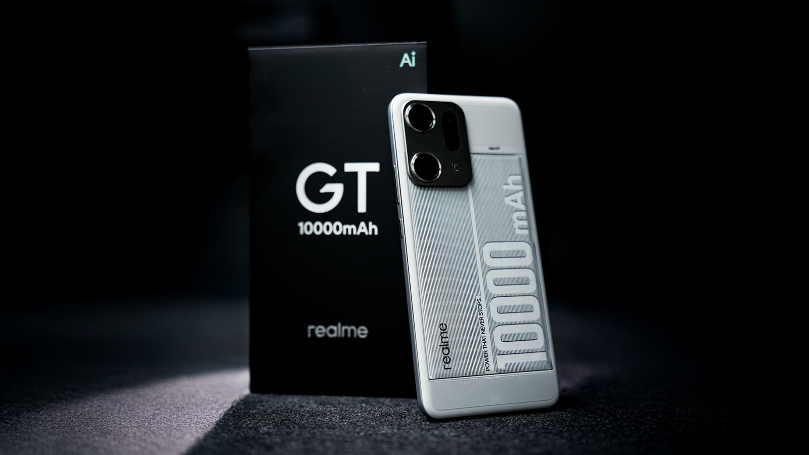 From Phonegram: A white realme GT smartphone is shown with '10,000 mAh' printed on its back, next to its black retail box on a dark surface, highlighting its impressive 10,000 mAh battery capacity.