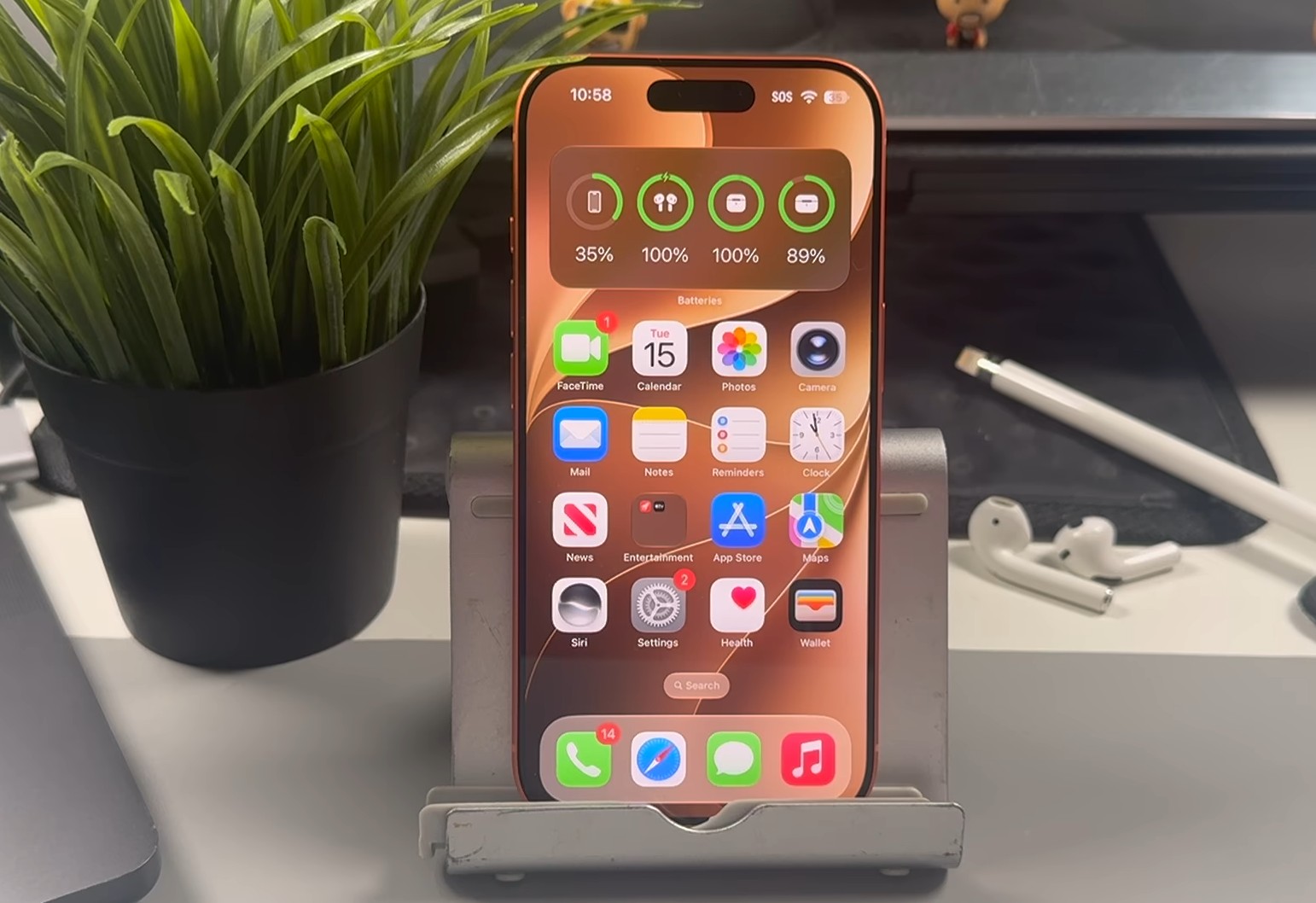 From the Phonegram website: An iPhone with a gold frame is placed on a stand, showing its home screen with the new «Siri» app in a prominent position. A potted plant, an Apple Pencil, and an AirPods case are on the desk in the background.