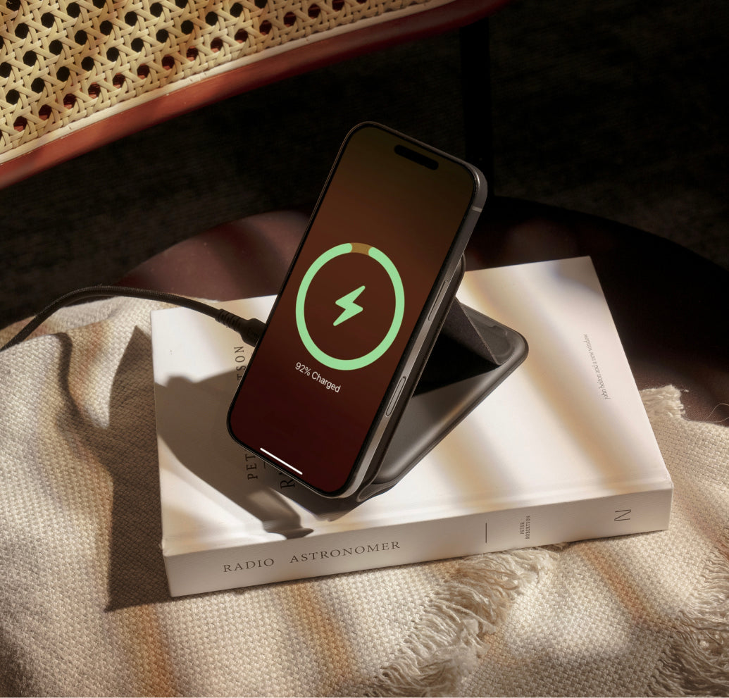من موقع فون إسلام: يظهر هاتف ذكي، يتم تحسين أداء بطارية آيفون أثناء شحنه على حامل لاسلكي، موضوعًا فوق كتاب مغلق بعنوان «عالم الفلك الراديوي»، مع كرسي منسوج وبطانية في الخلفية.