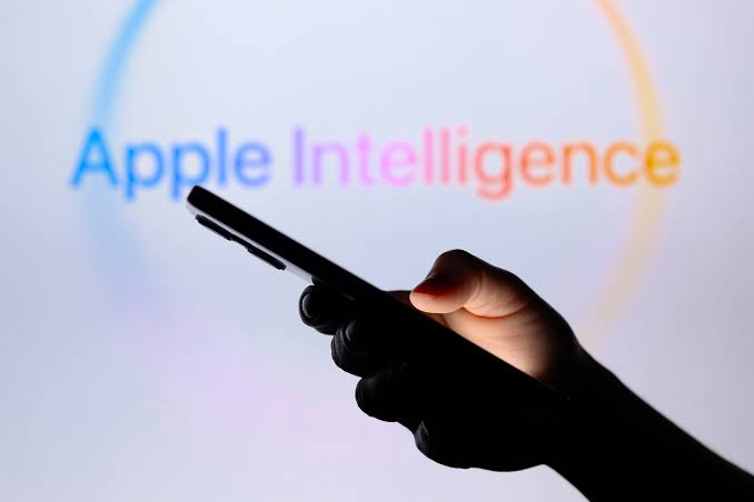 من موقع فون إسلام: يد تحمل هاتفًا ذكيًّا أمام شاشة تُعرض عليها شعار «Apple Intelligence»، المعروف باسم «ذكاء آبل».