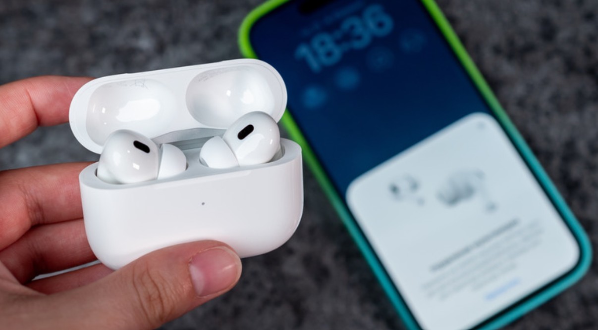 من موقع فون إسلام: يد تمسك بعلبة شحن بيضاء مفتوحة لسماعات AirPods، ويظهر في الخلفية هاتف ذكي يعرض شاشة اتصال لاستكشاف أسباب «الأصوات الغريبة» وإصلاحها.