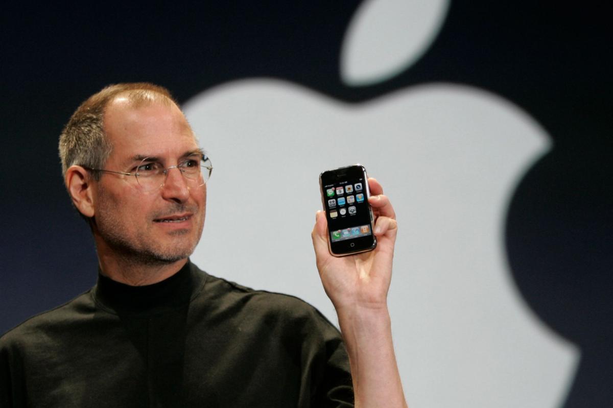 من موقع فون إسلام: ستيف جوبز يحمل جهاز آيفون من الطرازات الأولى يعمل بنظام التشغيل «iPhone OS 1» أمام شعار شركة آبل.