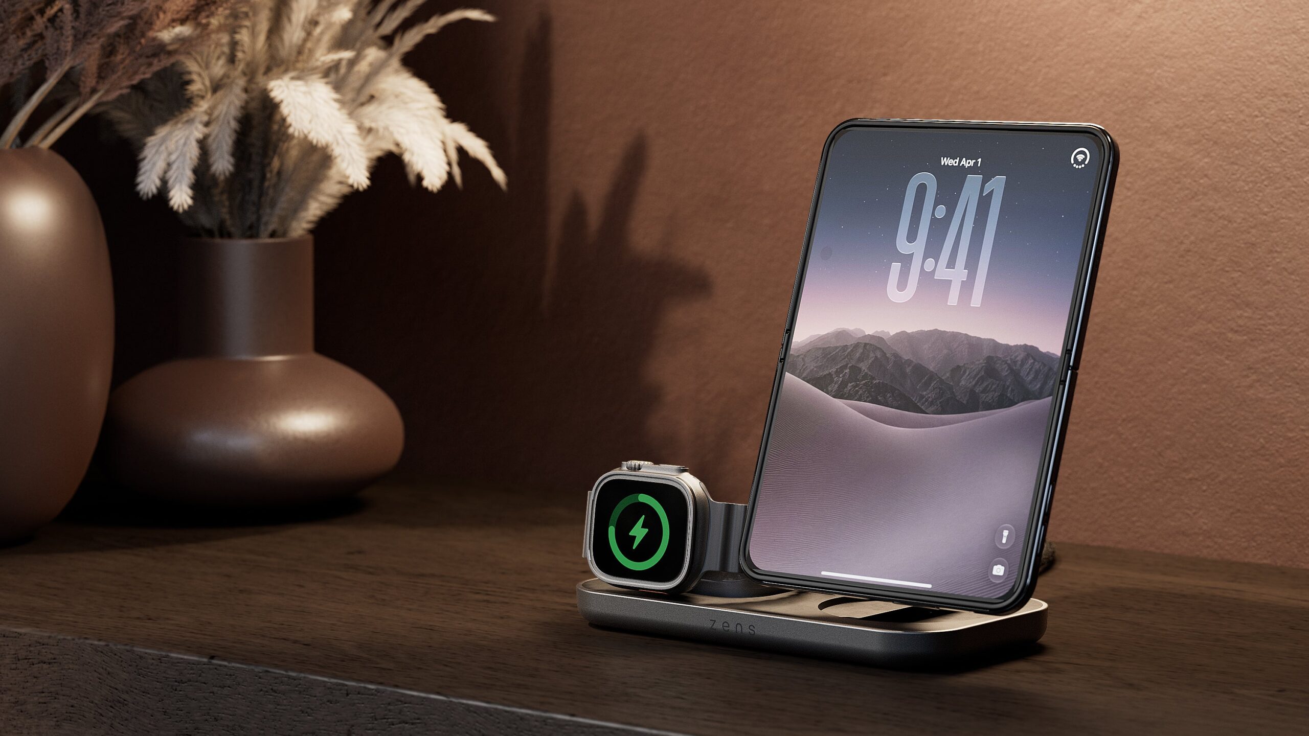من موقع فون إسلام: محطة شحن من طراز «Zens» مع ساعة ذكية وهاتف ذكي من طراز «iPhone Duo» مطوي، وكلاهما قيد الشحن على سطح خشبي.