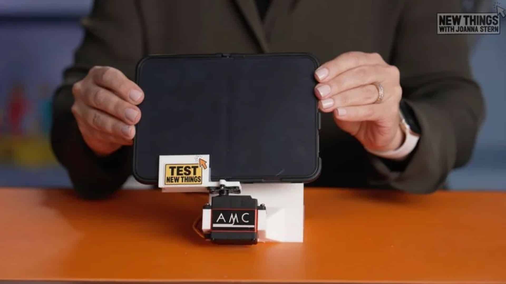 من موقع فون إسلام: يُمسك شخص ما بجهاز لوحي مُثبَّت على جهاز آلي من طراز AMC عليه ملصق كُتب عليه «TEST NEW THINGS»، مما يُشكّل إعدادًا مكونًا من شاشتين من طراز آي-فون ديو.