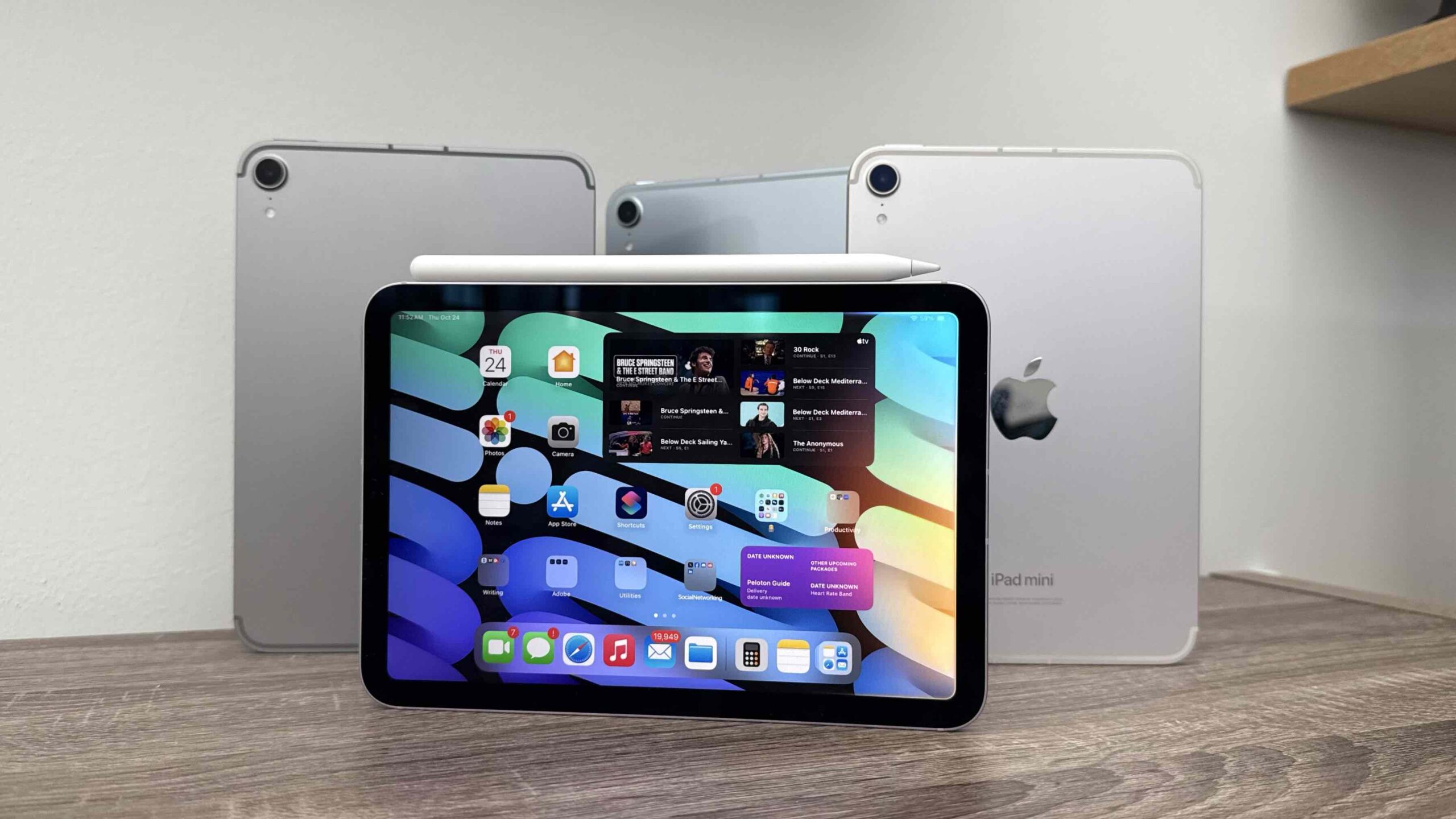 من موقع فون إسلام: جهاز iPad mini واحد بشاشة OLED يعرض شاشة رئيسية تحتوي على أيقونات التطبيقات والوحدات المصغرة، مع قلم Apple Pencil مثبت في الجزء العلوي، أمام جهازي iPad mini آخرين.